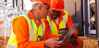 Two men wearing construction orange look at a laptop showing 3M training videos related to installing 3M electrical products.
